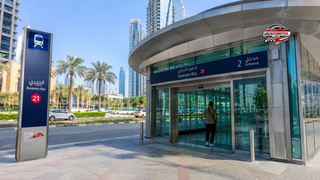 Realistic wide-angle view of Business Bay Metro Station Exit 2 in Dubai with modern skyscrapers, commuters walking outside the station, and Dubai Canal atmosphere during daytime.