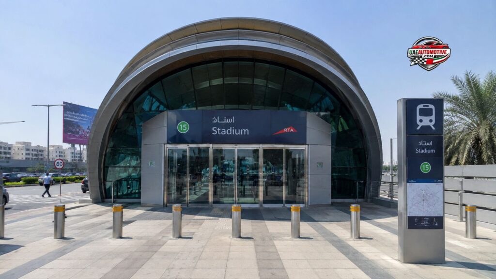 Front view of Stadium Metro Station in Dubai's Al Qusais district, featuring the station's iconic curved glass entrance, Metro signage, pedestrian plaza, and surrounding urban streetscape on a clear sunny day.